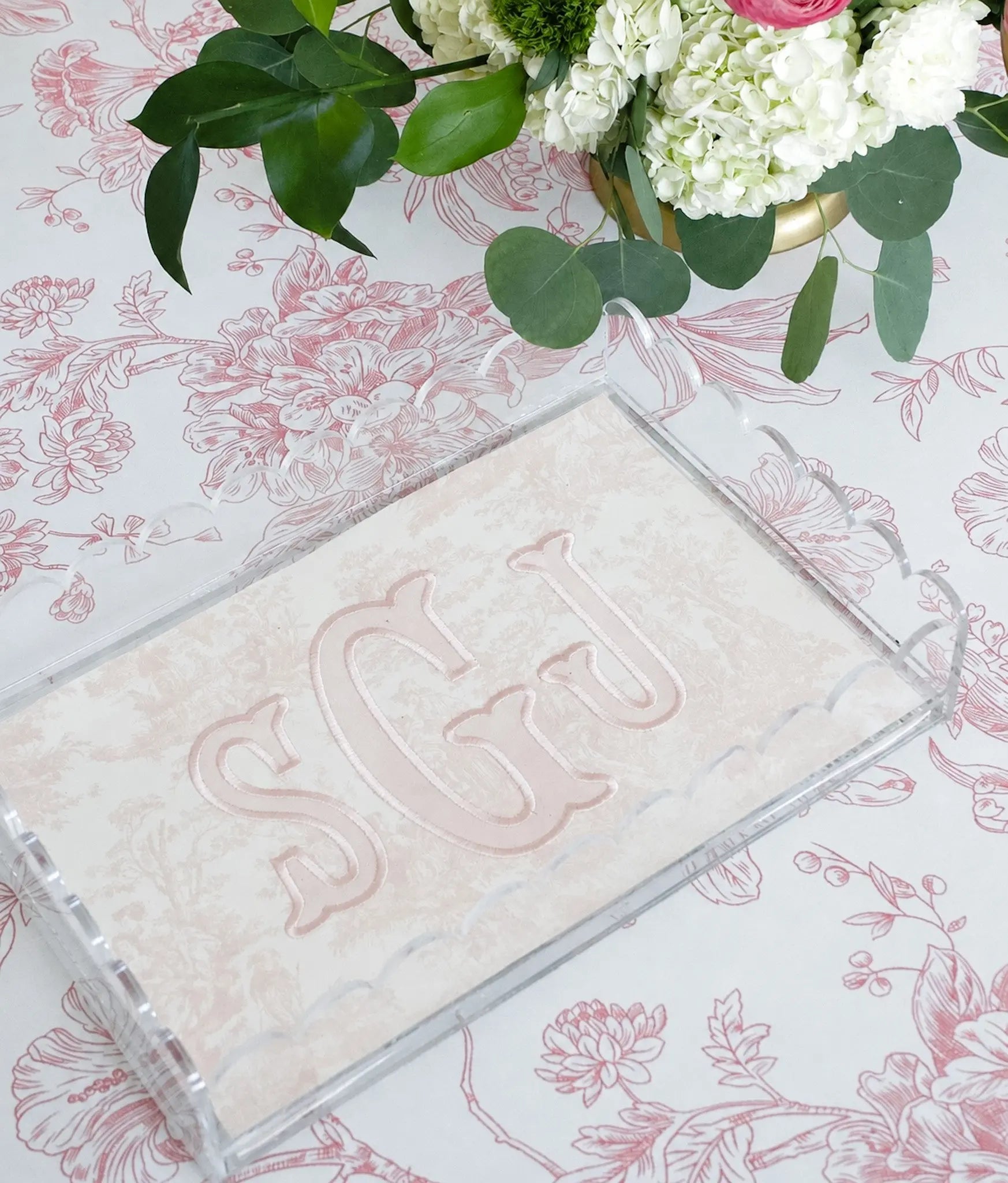 Pink toile monogram acrylic tray styled on a floral surface with green leaves