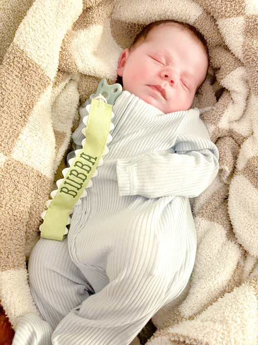 Personalized pacifier clip with embroidered baby name and decorative ribbon trim; styled view